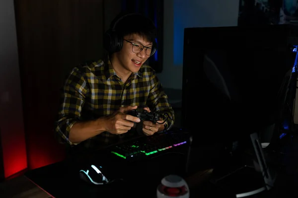 Handsome excited asian gamer guy in headphones enjoy and rejoicing while playing video games on computer in cozy room is lit with warm and neon light, gaming and technology e-sport concept.