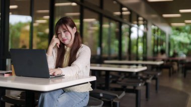 Genç Asyalı kadın açık kafede taşınabilir bilgisayar kullanıyor. O, esnek çalışma programı veya çevrimiçi çalışma programını yöneten uzak görevlere odaklanır.