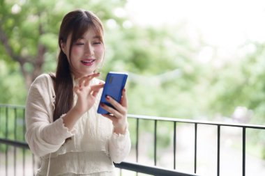 Rahatlamış Asyalı kadın gülümseyen yeşil arka planı bulanık balkonda akıllı telefon kullanıyor. Uzaktan çalışmanın tadını çıkarıyorum. Esnek yaşam tarzı..