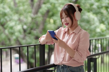 Genç Asyalı kadın dışarıda akıllı telefon ve kulaklık kullanarak dijital olarak bağlanıyor. Modern serbest yaşam tarzını ve uzaktan çalışma esnekliğini temsil eder.