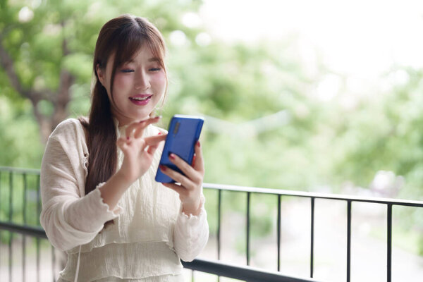 Расслабленная азиатка, улыбающаяся, использует смартфон на балконе с зеленым размытым фоном. Наслаждаясь гибким образом жизни удаленной работы.
