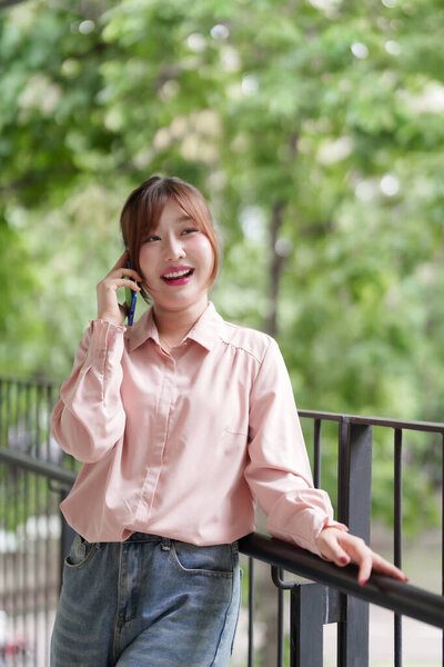 Счастливая взрослая азиатка разговаривает по телефону на улице. Она наслаждается гибкой дистанционной работой в окружении естественной зеленой среды.
