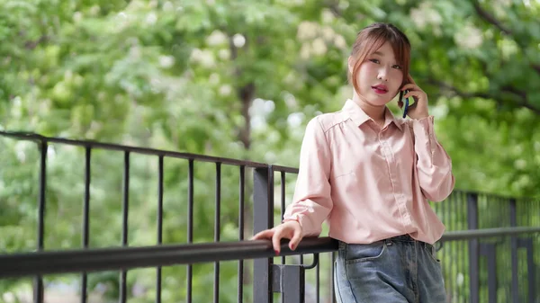 Genç Asyalı kadın iş için açık havada cep telefonu konuşuyor. Uzaktan kumandalı işçi esnek ve bağımsız yaşam tarzının tadını çıkarıyor. İletişim teknolojisi, doğayı güçlendiriyor.