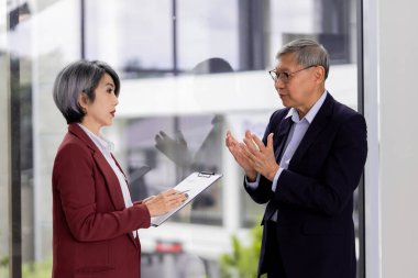 Adult Asian legal advisors collaborate in a modern office. Female lawyer consults older male client reviewing documents offering expert guidance.