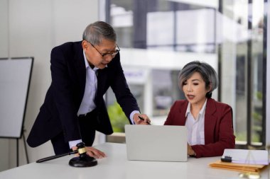 Asian legal experts collaborate on an important case using a laptop in a contemporary office setting. Two adult professionals review documents providing advice.