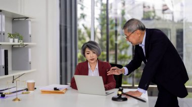 Two Asian legal professionals collaborate discussing important counsel. An experienced attorney offers guidance to a focused lawyer in modern office setting. Justice scales gavel symbolize law.