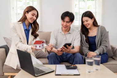Professional Asian agent offers expert home buying guidance to a happy young adult couple in their comfortable living space. Future property owners plan investments.