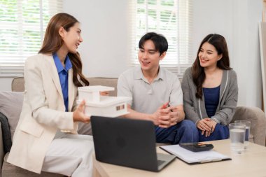Professional real estate agent explains house model to happy Asian couple during home visit. Discussing property investment and future home.