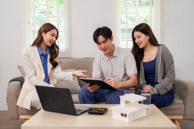 Professional Asian real estate agent consults clients at home. They discuss property investment and finalize buying agreement in modern comfort.