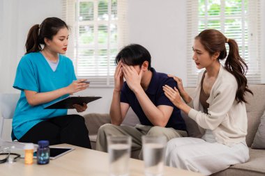 Empathetic healthcare provider counsels adult Asian man at home addressing emotional distress while supportive partner offers comfort. Focus on wellness.