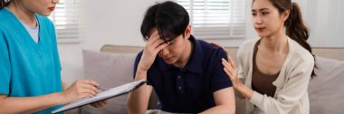 An Asian adult man expresses distress during a home mental health session. His partner offers comfort while a therapist provides guidance and support for wellbeing.