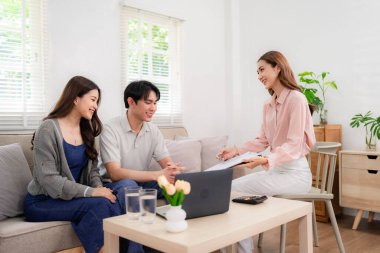 Professional Asian agent discusses vital health insurance plans with a happy young couple in their comfortable living room Expert guidance for future financial security