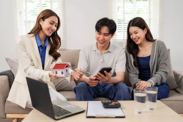 Professional Asian agent offers expert home buying guidance to a happy young adult couple in their comfortable living space. Future property owners plan investments.