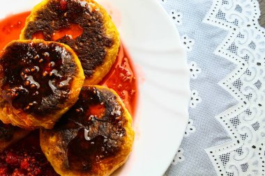 Koyu kahverengi bir masaüzerinde, beyaz desenli bir masa örtüsü, yiyecek bir tabak vardır. Ahududu reçeli ile Lor cheesecakes