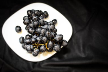 Yatay fotografın siyah arka planında beyaz bir plaka üzerinde bir dalda taze koyu üzümler uzanıyor.