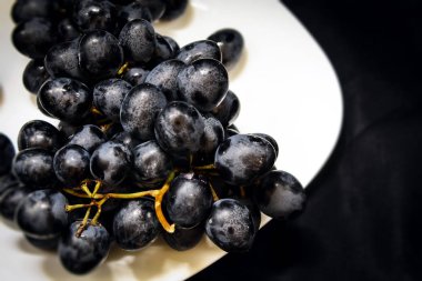 Yatay fotografın siyah arka planında beyaz bir tabakta taze koyu üzümler yatıyor.