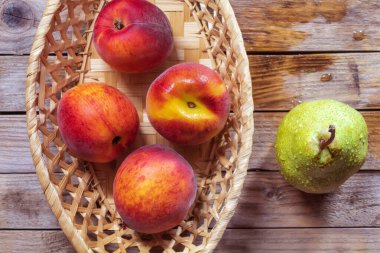 Yatay fotoğraf taze ve olgun meyveleri gösteriyor. Sepette dört şeftali var, yanında da bir damla su olan sarı bir armut. Ahşap zemine yerleştirilmiş meyveler