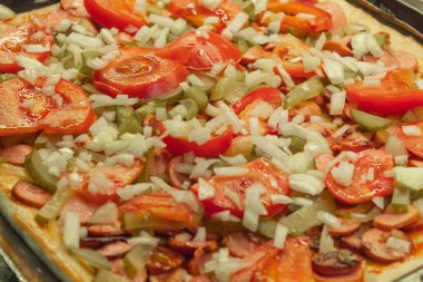 Yatay resim ev yapımı pizzayı gösteriyor, yakında fırına konacak. İnce bir hamur tabakasında taze sebze, sosis ve ketçap var..