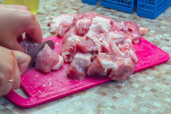 Pictured is an image of the process of cooking dinner from meat. On a pink plastic cutting board is fresh meat of the animal, cut into small pieces. You can see the hands of the man who cuts the meat