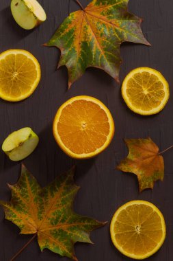 Dikey fotoğraf taze meyve ve üç akçaağaç yaprağı bileşimini gösteriyor. İki yaprak büyük, biri küçük. Portakalın ortasında ve kenarlarında üç tane yuvarlak dilim var. Bir de iki dilim elma.