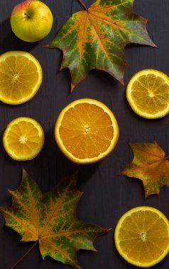 Dikey fotoğraf taze meyve ve üç akçaağaç yaprağı bileşimini gösteriyor. İki büyük yaprak, bir küçük. Ortasında yarım portakal, ve kenarlarında dört yuvarlak dilim var. Bir de küçük bir elma var.