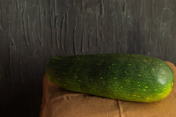 In the horizontal photo, a whole green zucchini is lying on a kitchen towel. Zucchini, raw, with rind for cooking