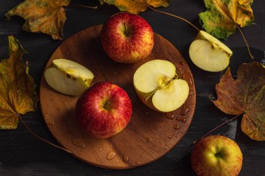 Yatay bir fotoğraf tahtada duran taze kırmızı elmaları gösteriyor. Etrafta kuru akçaağaç yaprakları var. Üç tam elma ve üç dilim hamurlu ve tohumlu. Nesneler siyah ahşap bir arka planda yatar