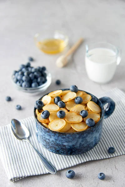 Blue Bowl of mini pancake cereal with blueberries, maple syrup and milk on concrete background. Tiny pancakes, new food trend concept. Cute healthy breakfast or snack. Recipe, menu.