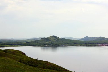 Panoramik manzara. Görkemli dağların ortasında sisli, temiz bir dağ gölü olan bir göl. Yeşil çimenler. Gölün çevresindeki daireler.