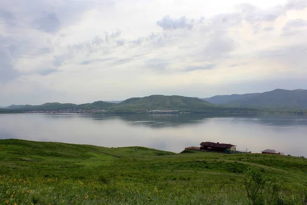 Panoramic view. A lagoon with a clean mountain lake in the midst of majestic mountains in a haze of fog. Green grass. Apartments around the lake — Stock Photo, Image