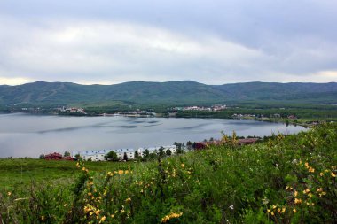 Panoramik manzara. Görkemli dağların ortasında sisli, temiz bir dağ gölü olan bir göl. Yeşil çimenler. Gölün çevresindeki daireler.