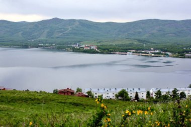 Panoramik manzara. Görkemli dağların ortasında sisli, temiz bir dağ gölü olan bir göl. Yeşil çimenler. Gölün çevresindeki daireler.