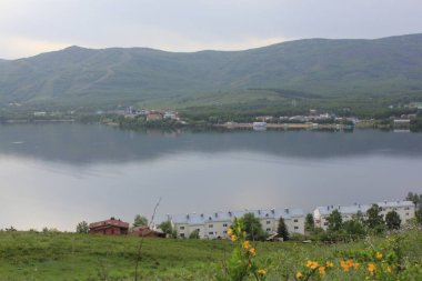 Panoramik manzara. Görkemli dağların ortasında sisli, temiz bir dağ gölü olan bir göl. Yeşil çimenler. Gölün çevresindeki daireler.