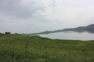 Panoramik manzara. Görkemli dağların ortasında sisli, temiz bir dağ gölü olan bir göl. Yeşil çimenler. Gölün çevresindeki daireler.
