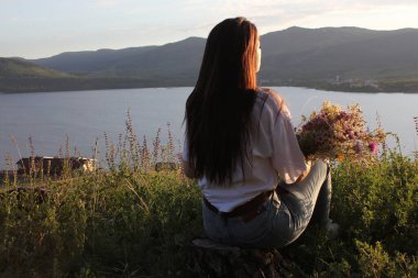 Elinde bir buket çiçekle genç bir kız bir dağ gölünün önündeki dağın tepesindeki günbatımına hayran. Gün batımı