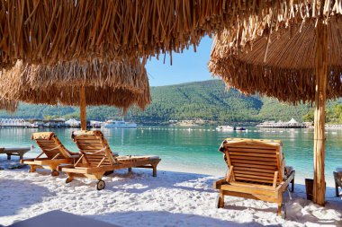 Zümrüt suyu, yatlar ve arka planda yeşil tepeler olan beyaz bir sahilde bambu şemsiyeler. Mutlu Lüks Turizm Gebeliği