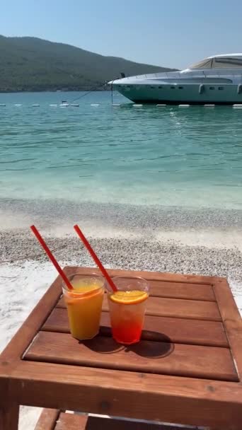 4K-Video Zwei Gläser einer Cocktailaperole mit Orangenscheiben stehen auf einem Holzständer an einem schneeweißen Strand vor dem Hintergrund einer schneeweißen Jacht im smaragdgrünen Meer. Glücklicher Lebensstil — Stockvideo