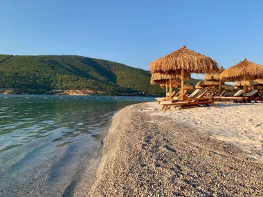 Beyaz kum, bambu palmiyeleri ve güneş panjurlarıyla zümrüt Ege sahilinin şık manzarası. Lüks rahatlama konsepti. Arkasında dağ olan beyaz bir sahil.