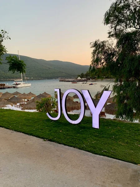 Large letters: JOY, Panoramic view of the emerald Aegean coast with white sand, bamboo palms and sun loungers. Luxury leisure relaxation concept. White beach with a mountain in the background — Stock Photo, Image