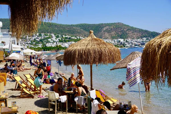 Бодрум, Турция - август 2020 года: Много людей на пляже залива Кумбахче. Бодрум, провинция Мугла, Турция. Люди загорают, обедают в кафе, гуляют по пляжу. Стоковая Картинка