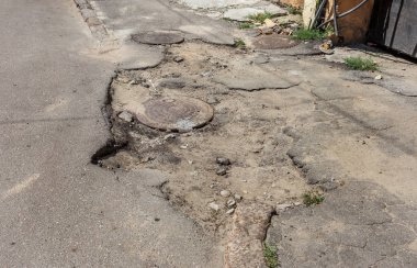Damaged bad asphalt road, cracked asphalt with potholes and stains. A very bad asphalt road with large holes, a sewer hatch in the pit of the road. Numerous dangerous failures clipart