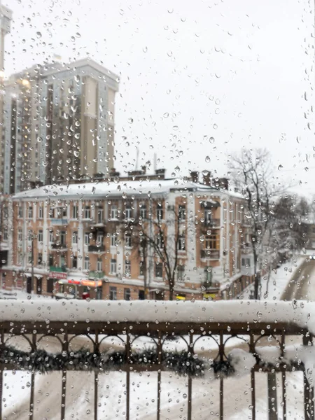 Drops of water on the glass. Snow melting. From the wet snow on the window glass formed a drop of water, flowing down. Winter landscape of the snow-covered city outside the window.