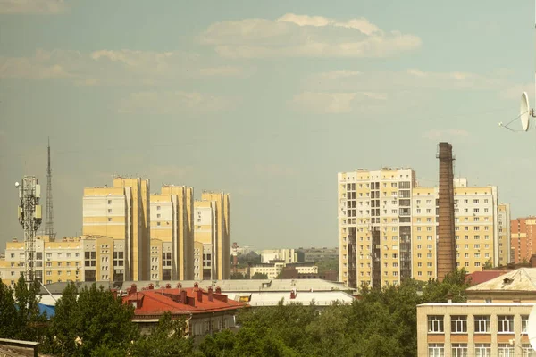 Şehirdeki pencereden kent manzarası: yüksek binalar, çatılar ve fayanslar, baz istasyonları, televizyon antenleri ve arka planda bulutlar olan büyük sanayi boruları