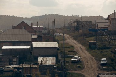 Ahşap evler, fayanslar, arabalar ve arka planda dağ aralığı ve tepesi olan toprak zemin.