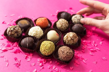 Kadın elinde şekerlemeyle pembe arka planda farklı çikolatalı pralinler tutuyor. 