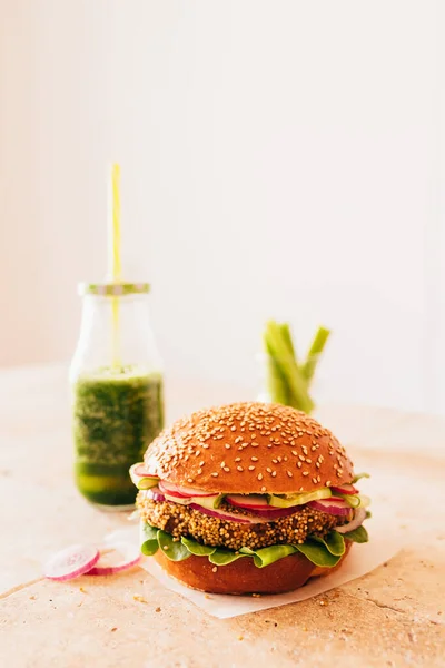 Bir şişe yeşil smoothie ile vejetaryen burger ve bardakta kerevizli havuç. Taş bej masa