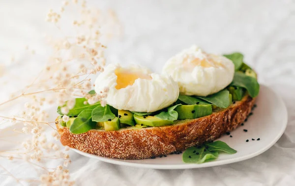Tam tahıllı ekmek, avokado, roka ve küçük çingene çiçekleriyle süslenmiş nigella serpiştirilmiş haşlanmış yumurtalı sandviç. Tekstil ve bej arka plan. Boşluğu kopyala