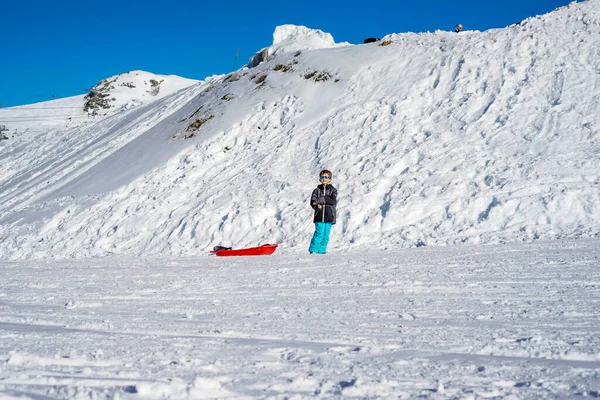 Jongen met rode slee in de sneeuw. Sneeuwberg achtergrond. — Stockfoto