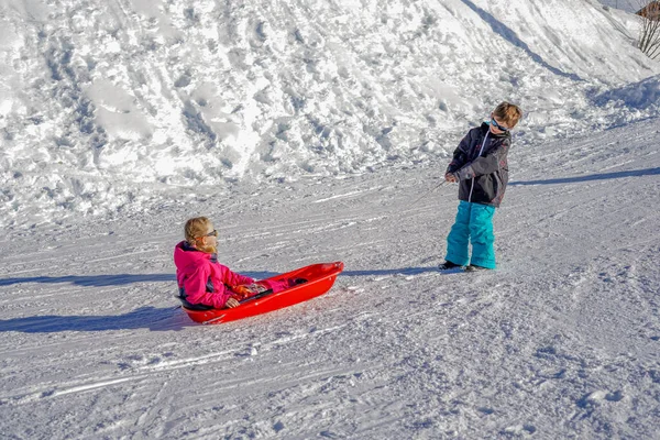 Ο αδερφός τραβούσε τα έλκηθρα των αδελφών του. Κοριτσάκι και αγόρι απολαμβάνουν τη βόλτα με έλκηθρο. Παιδικό έλκηθρο. Τα παιδιά παίζουν έξω στο χιόνι. — Φωτογραφία Αρχείου