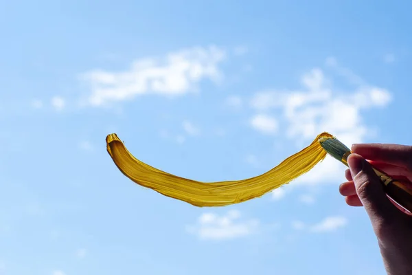 Pintura femenina de primer plano con un pincel una sonrisa amarilla en la ventana, cielo azul. El concepto de alegría, felicidad, salud —  Fotos de Stock